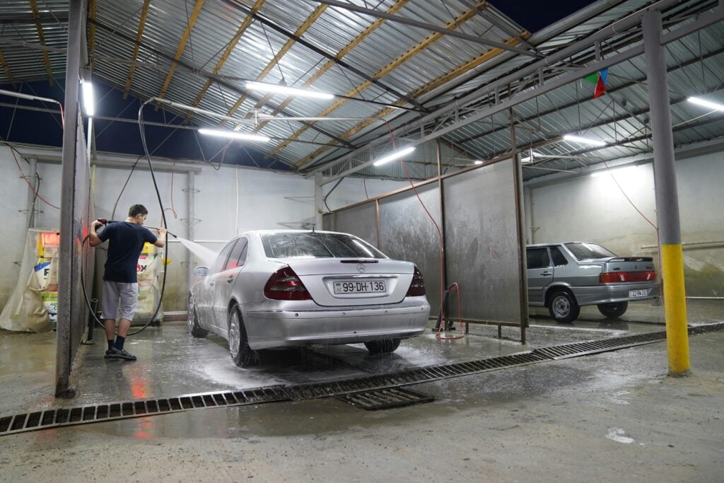 A man uses a pressure washer to clean a silver sedan in a covered car wash at night.